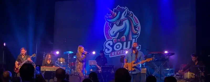 Le groupe complet sur scène : chanteuse, bassiste, guitariste, claviers et batteries, devant un grand écran avec le logo de la licorne Soul Revenge.